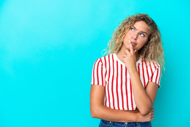 Chica con el pelo rizado aislado sobre fondo azul teniendo dudas mientras mira hacia arriba