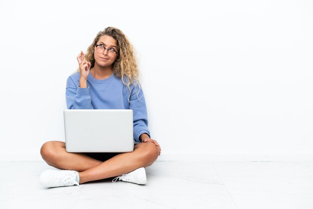 Chica con el pelo rizado con una computadora portátil sentada en el suelo con los dedos cruzados y deseando lo mejor