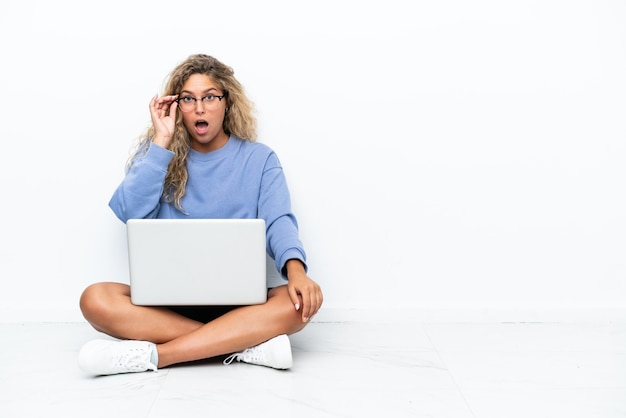 Foto chica con el pelo rizado con una laptop sentada en el suelo con gafas y sorprendida