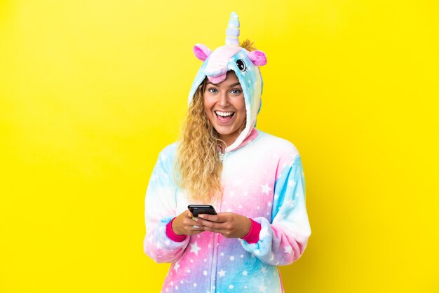 Chica con el pelo rizado con un pijama de unicornio aislado en un fondo amarillo sorprendida y enviando un mensaje