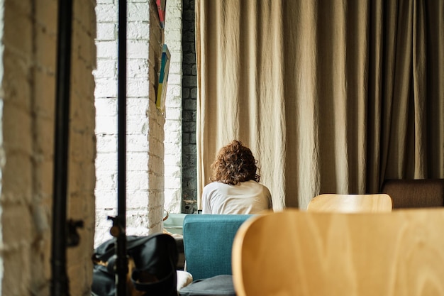 La chica con el pelo rizado se sienta sola con la espalda en el café y trabaja en una cortina beige de fondo Copiar espacio