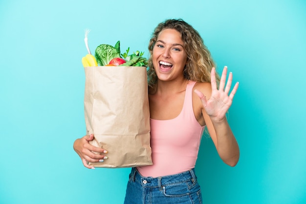Chica con el pelo rizado sosteniendo una bolsa de compras aislada en un fondo verde contando cinco con los dedos