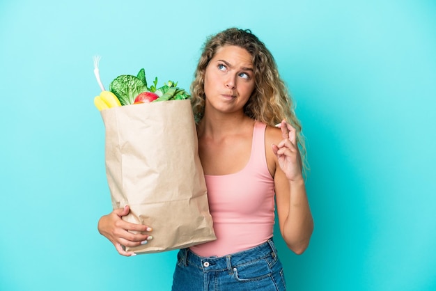 Chica con el pelo rizado sosteniendo una bolsa de compras aislada en un fondo verde con los dedos cruzados y deseando lo mejor