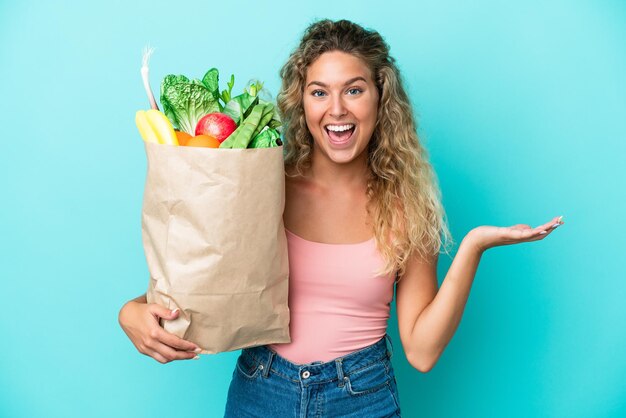 Chica con el pelo rizado sosteniendo una bolsa de compras aislada en un fondo verde con una expresión facial sorprendida