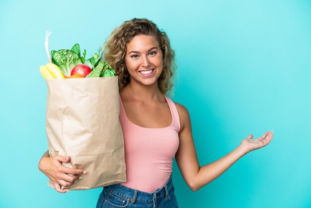 Chica con el pelo rizado sosteniendo una bolsa de compras aislada en un fondo verde extendiendo las manos a un lado para invitar a venir