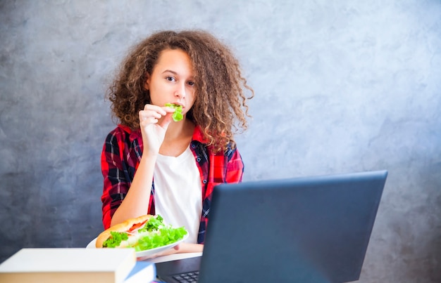 Foto chica de pelo rizado trabajando en la computadora portátil y comiendo sandwich