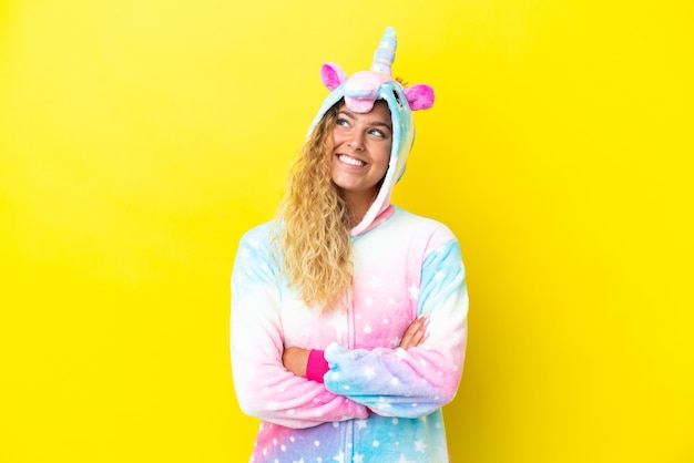 Chica con el pelo rizado vistiendo un pijama de unicornio aislado sobre fondo amarillo mirando hacia arriba mientras sonríe
