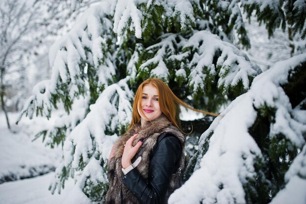 Chica de pelo rojo en abrigo de piel caminando en el parque cubierto de nieve de invierno.