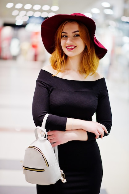 Chica de pelo rojo de moda en vestido negro y sombrero rojo con mochila de mujer en el centro comercial.