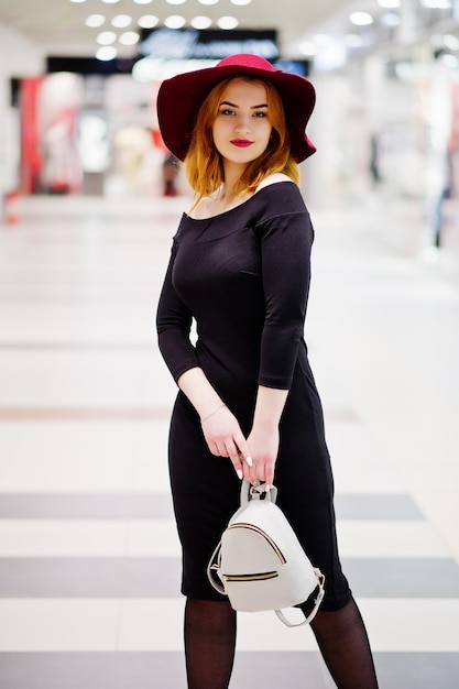 Chica de pelo rojo de moda en vestido negro y sombrero rojo con mochila de mujer en el centro comercial.