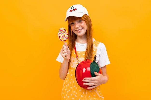 Chica con el pelo rojo posando en la cámara con una pequeña bolsa de manzana come un lolipop aislado en amarillo