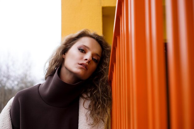 Una chica con el pelo rojo y rizado con una bata blanca posa en las escaleras de estacionamiento de color amarillo anaranjado. Estilo de ciudad - Urbano