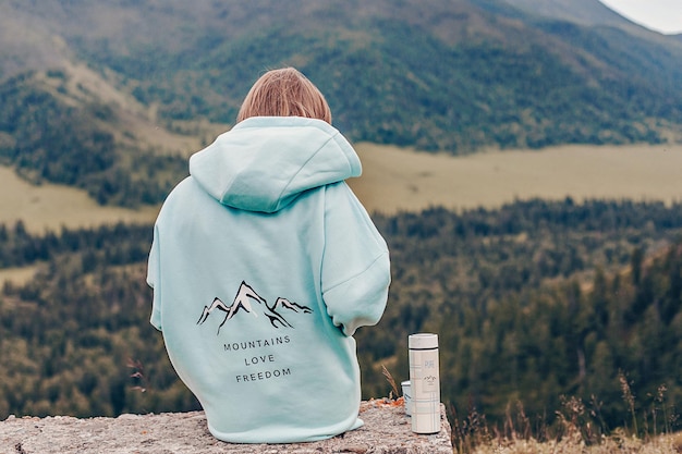 Una chica con el pelo rubio está sentada con la espalda apoyada en una piedra con una sudadera con capucha azul con un estampado. Hay un termo y una taza de viajero cerca. Fondo de una vista de la montaña verde.