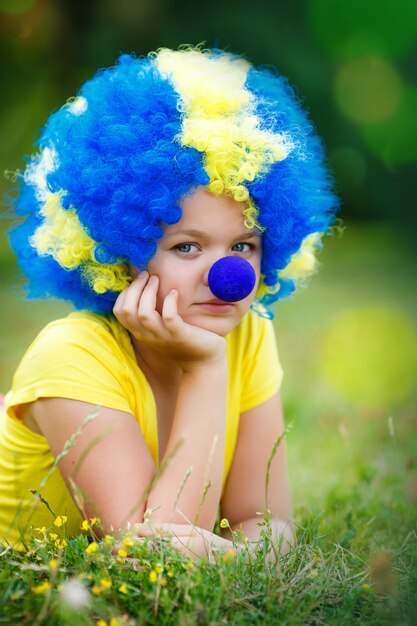 Chica con peluca de payaso con nariz azul está tumbado en la hierba verde en el parque