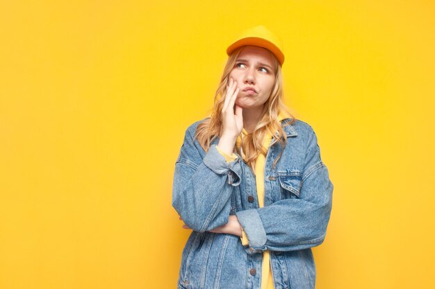 Una chica pensativa con una chaqueta vaquera y una gorra se para sobre un fondo amarillo y piensa