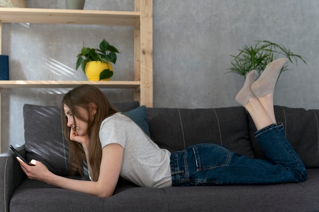 Chica pensativa se encuentra en el sofá con el teléfono en las manos. Adicción al teléfono entre los jóvenes jóvenes.