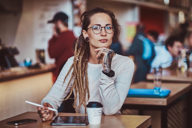 Una chica pensativa con estilo está dibujando en un bloc de notas digital mientras está sentada en un café con una taza de café.