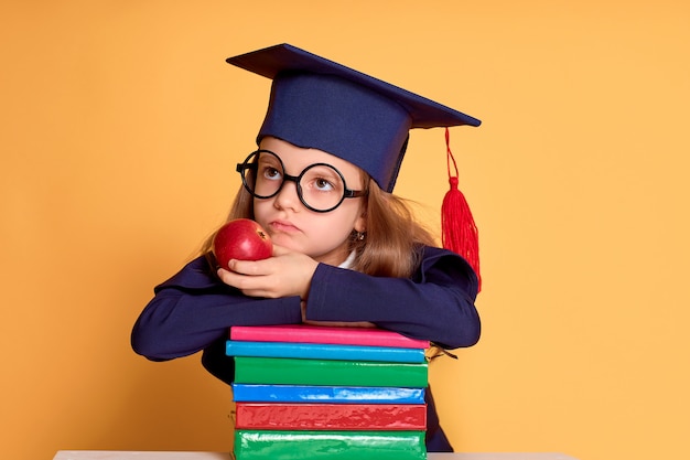 Chica pensativa con gafas y ropa de graduación pensando mientras se acuesta en los coloridos libros