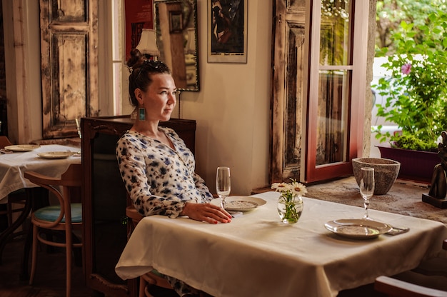 Chica pensativa en un vestido de verano está descansando en una mesa en un café cerca de la ventana