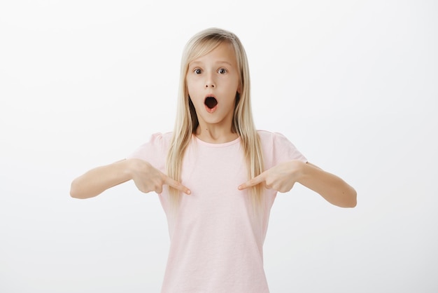 Chica perdiendo el habla del asombro. Retrato de una linda hija sorprendida sorprendida con cabello rubio, apuntando al pecho con los dedos índices