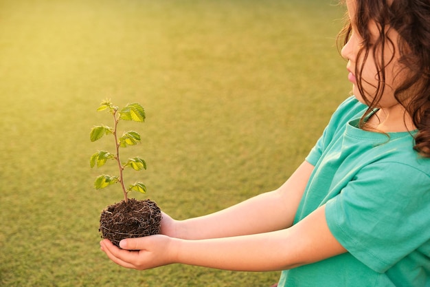 Chica de perfil con el pelo largo y rizado con camiseta verde con las manos extendidas sosteniendo una pequeña planta verde que está brotando sobre un fondo de hierba verde desenfocado