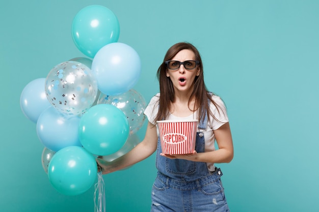 Una chica perpleja con gafas 3d viendo una película sostiene un cubo de palomitas de maíz celebrando con coloridos globos de aire aislados en un fondo azul turquesa. Emociones de fiesta de cumpleaños en el concepto de cine.