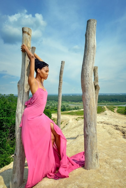 La chica de pertenencia africana con un vestido rosa se encuentra en una colina alta apoyada contra un árbol seco