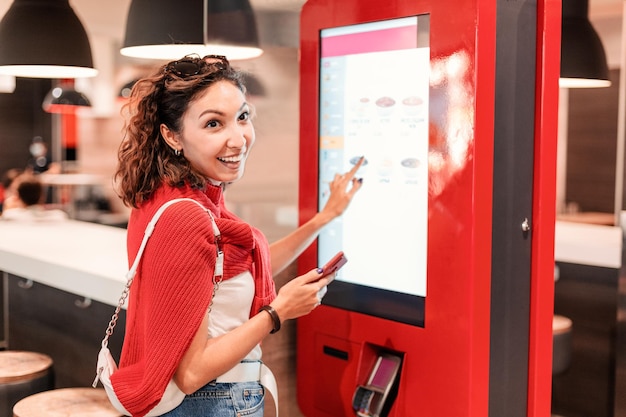 Una chica pide comida y almuerzo en un restaurante de comida rápida usando un quiosco de autoservicio o una terminal con una pantalla Equipamiento de comercio moderno