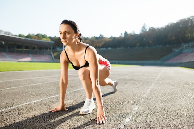 Chica de pie en el punto de inicio en el estadio antes de la pista de atletismo. Mujer joven en top negro, pantalones cortos rosas y zapatillas blancas listas para correr. Al aire libre, deporte