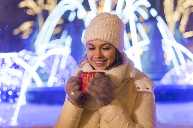 Chica de pie sobre la nieve de fondo de la ciudad de Navidad de invierno, se encuentra un sombrero de chaqueta caliente, sostiene una taza de té con bebidas de té caliente.
