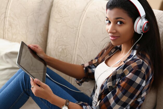 Una chica de piel oscura con auriculares con tableta se sienta en el sofá