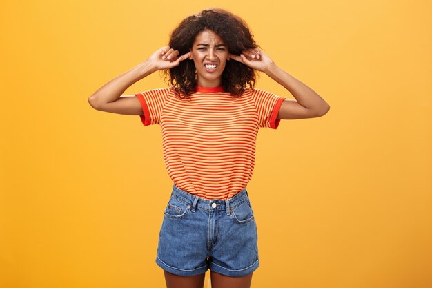 Chica de piel oscura disgustada con el pelo rizado frunciendo el ceño apretando los dientes por la incomodidad cerrando las orejas con los dedos índices sobre la pared naranja