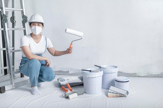 Foto chica pinta una pared blanca con un rodillo reparación del interior joven decoradora pintando una pared en la habitación vacía constructora de conceptos o pintora en casco con rodillo de pintura sobre la habitación vacía