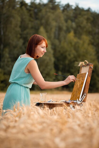 Chica pintora profesional trabajando en una pintura en el campo de trigo maduro. Artista tranquilo y alegre terminando el dibujo, foto vertical.