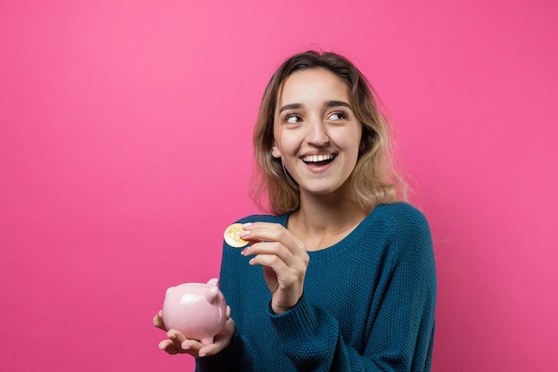 Foto chica pone en la alcancía bitcoin físico joven sobre fondo rosa sosteniendo alcancía y se regocija gesticulando el concepto de confiabilidad de las inversiones en efectivo y seguros