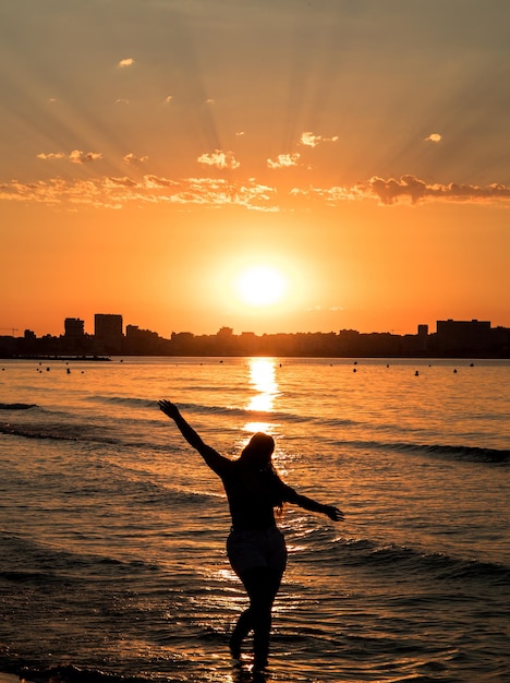 Chica posando con los brazos en alto en un amanecer en la playa del postiguet en Alicante, con edificios contra