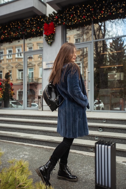 Chica posando en la carretera en el fondo de invierno Mujer joven divertida y glamorosa con una sonrisa con un elegante abrigo de piel largo azul Concepto de piel y moda