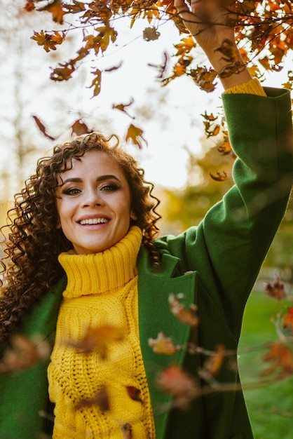 Una chica positiva con una hermosa sonrisa de dientes blancos con ropa cálida de otoño se para debajo de un árbol con hojas amarillas y las toca con la mano