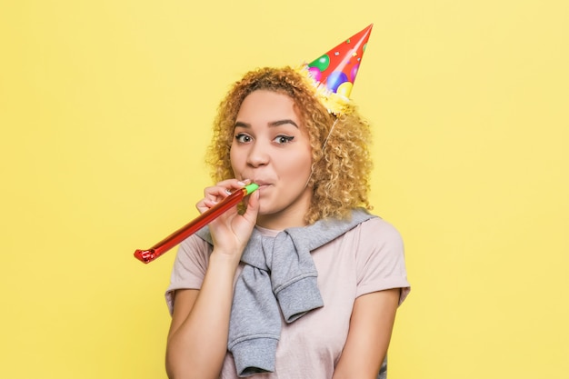 La chica positiva sostiene un silbato de cumpleaños y le sopla un poco de pelo. También tiene un sombrero de cumpleaños en la cabeza.