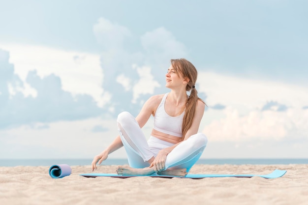 Chica practicando yoga en la mañana en el fondo del mar Joven mujer caucásica se sienta en la estera de yoga en la playa