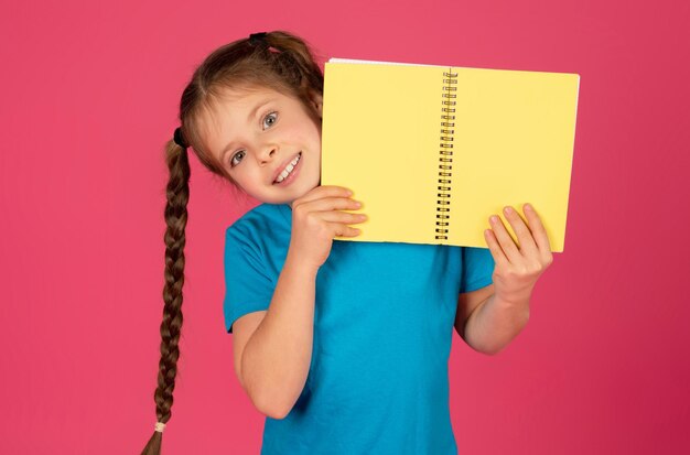 Foto chica preadolescente feliz con trenzas largas escondida detrás de un bloc de notas brillante