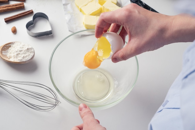 Chica preparando galletas en la cocina, rompiendo un huevo, primer plano. Una mujer prepara masa para deliciosos pasteles. Instrucciones paso a paso. Mezcla de diferentes ingredientes para la masa. Fondo.