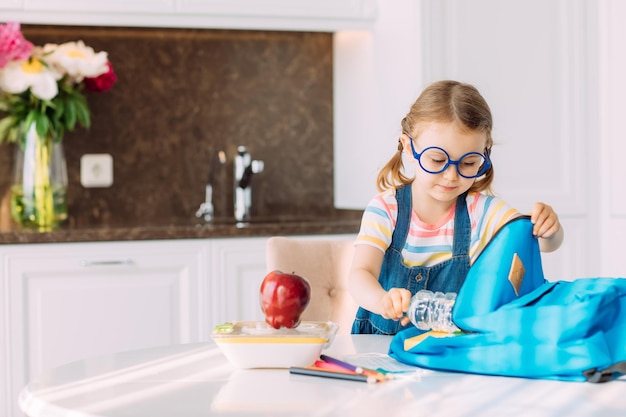 Chica preparando mochila para la escuela en casa en la mesa en la mañana soleada cocinaEducaciónaprendizaje