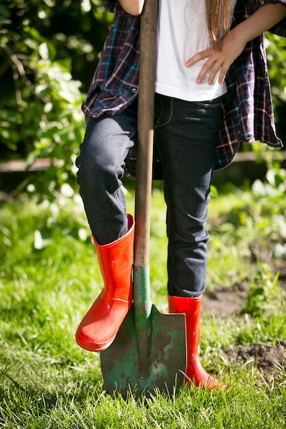 Chica de primer plano en botas de goma roja sosteniendo la pierna en la pala en el jardín