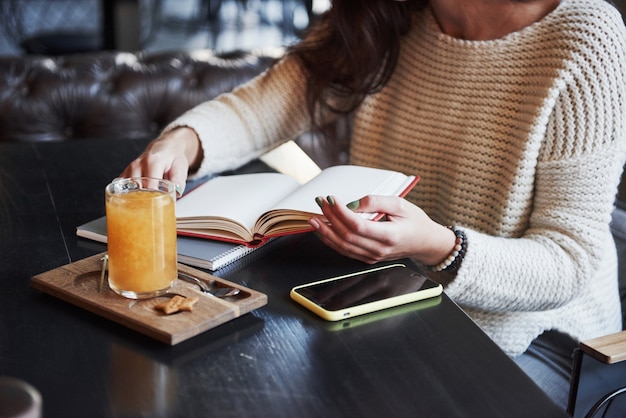 Chica en proceso de estudio. Recorta la foto de una mujer que lee el libro en el restaurante con una bebida amarilla y un teléfono inteligente en la mesa