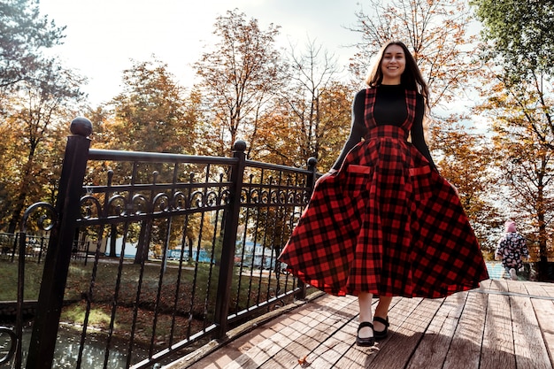 Chica en puente con vestido largo escocés rojo Chica con cabello largo y castaño en el parque otoño