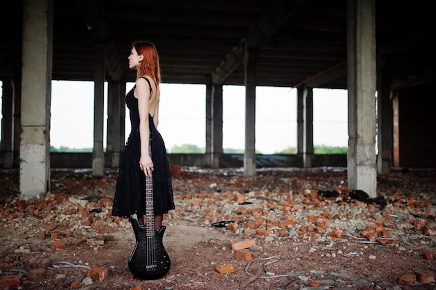 Chica punk pelirroja vestida de negro con bajo en un lugar abandonado Retrato de mujer gótica músico