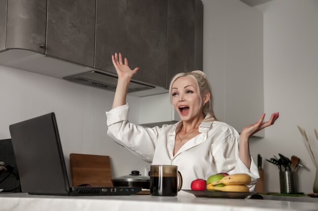 Chica que trabaja en la computadora portátil en la cocina en casa y muy emocional. Gestos de mujer con las manos. Negocios desde casa. Linda mujer eslava en la cocina mira la computadora. Emociones femeninas. Concepto de tarea