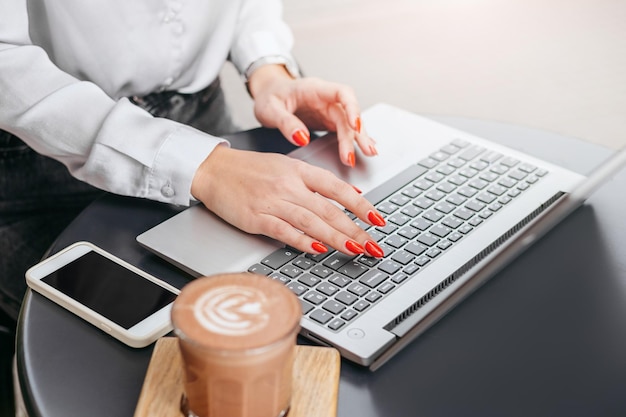 Chica que trabaja con una computadora portátil y un teléfono móvil sentada en una mesa en un café