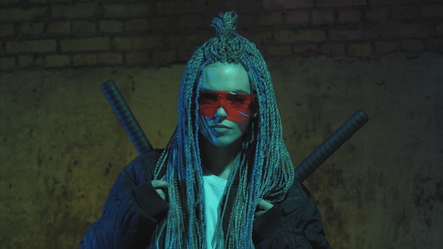 Chica con rastas y katanas en gafas rojas posando contra una pared de ladrillos de neón